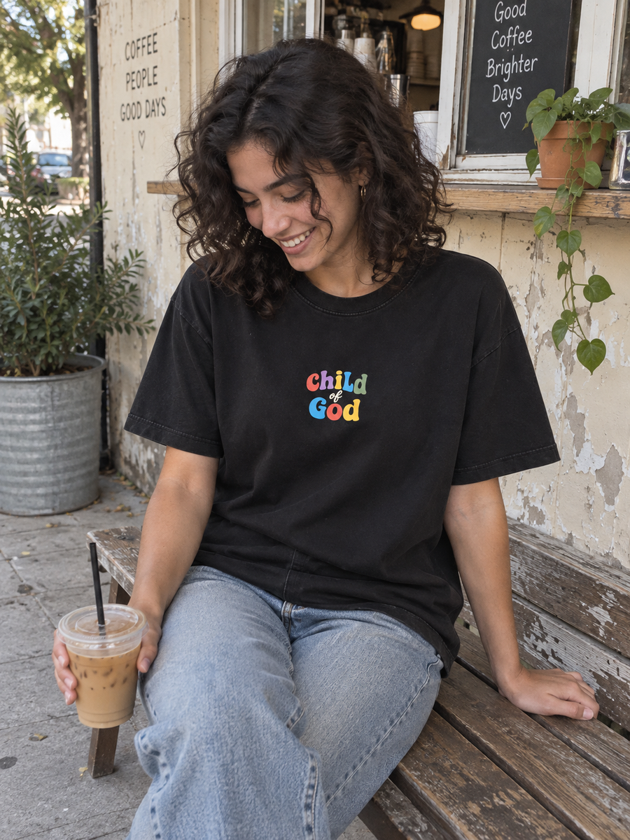 Woman wearing the Child of God front print, enjoying coffee on a bench outside a neighborhood kiosk.