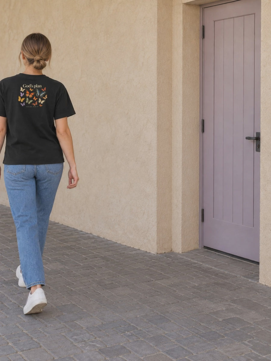 God’s Plan Tee — one woman walking naturally into frame, back butterfly print, lavender-door stucco passage