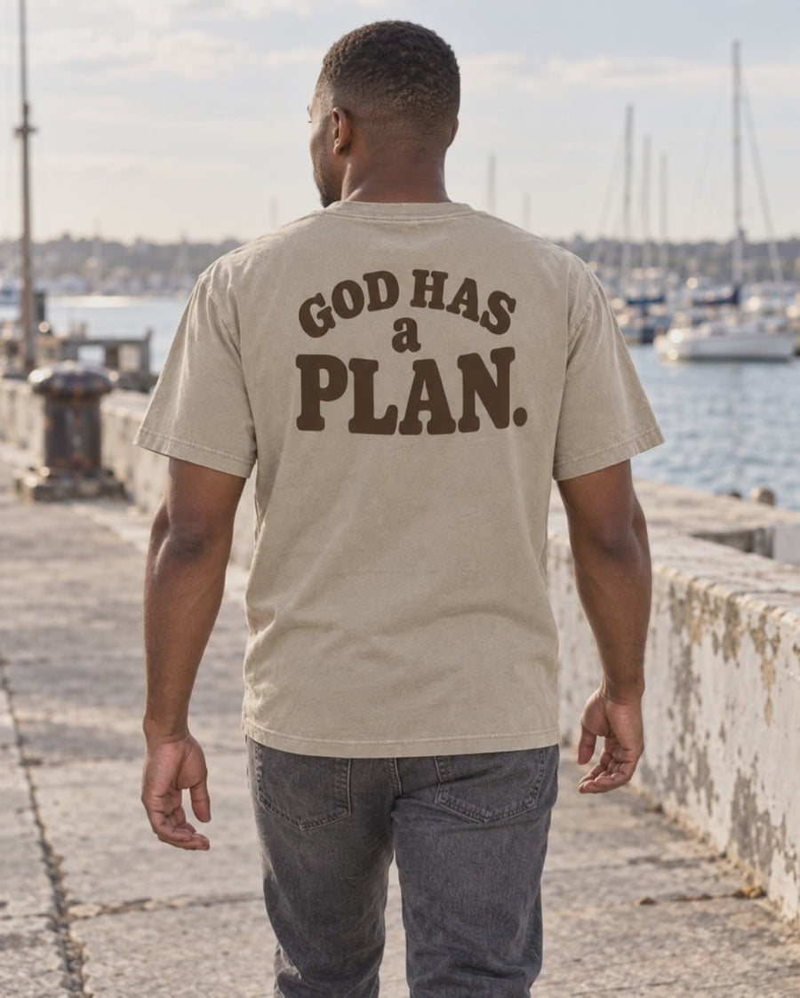 Khaki God Has a Plan tee: new male model walking along a harbor promenade, then new female model beside a weathered coastal wall; back and front lettering views.