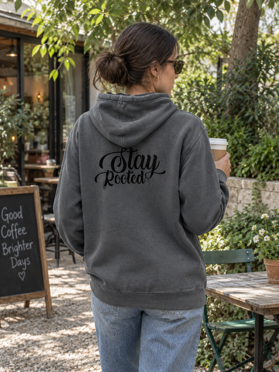 Stay Rooted Unisex Pigment-Dyed Hoodie Sweatshirt — Woman holding coffee in leafy courtyard, back print visible