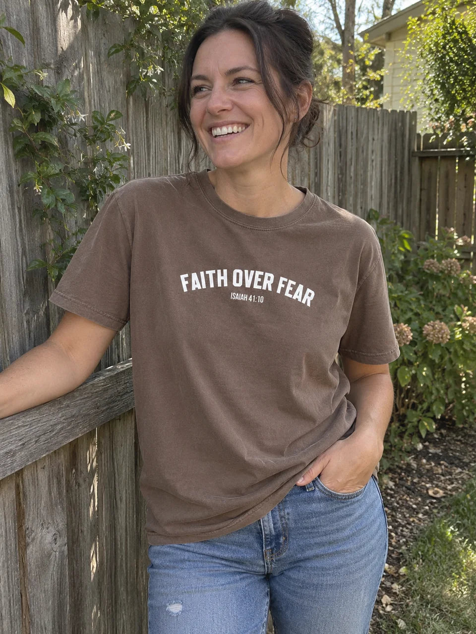 Brown Faith Over Fear tee styled with jeans beside a garden fence
